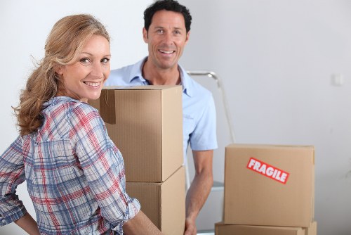 A smiling woman with light brown, wavy hair dressed in a casual checked shirt is holding a medium-sized cardboard box in a residential interior, likely a house or flat in Shoreditch, preparing for a move. Behind her, a man with short dark hair wearing a light blue shirt is standing near a stack of two additional cardboard boxes, one of which is marked with a red